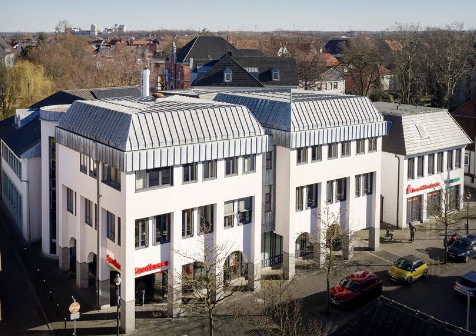 Außenansicht eines modernen mehrstöckigen Bürogebäudes mit heller Fassade und silbernem Dach. Im Erdgeschoss befindet sich eine Sparkassen-Filiale. Autos und Fußgänger sind auf der Straße davor zu sehen.