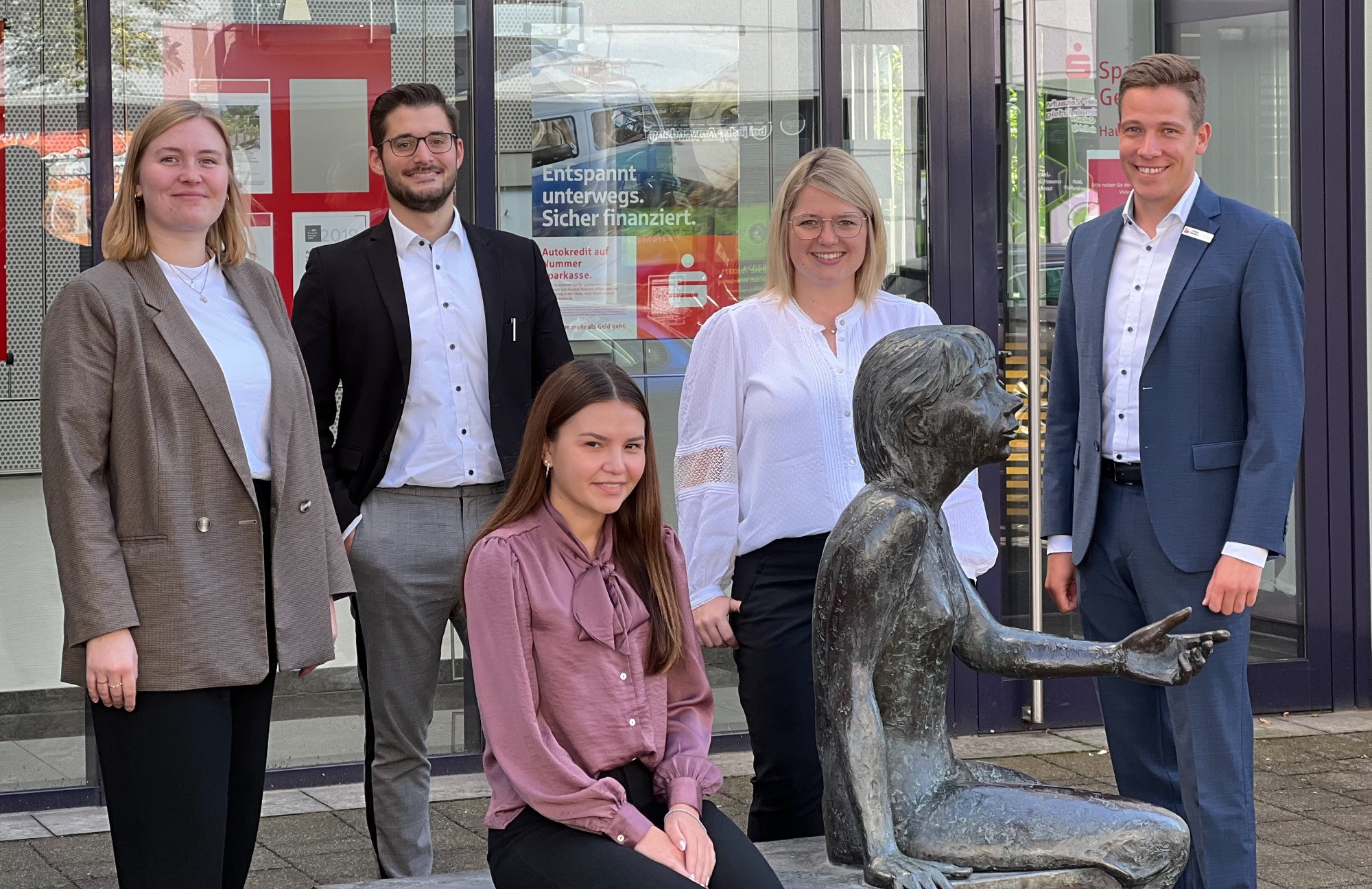 Fünf junge Erwachsene stehen und sitzen vor einem Gebäude der Sparkasse. Sie posieren lächelnd um eine bronzene Sitzstatue im Vordergrund.