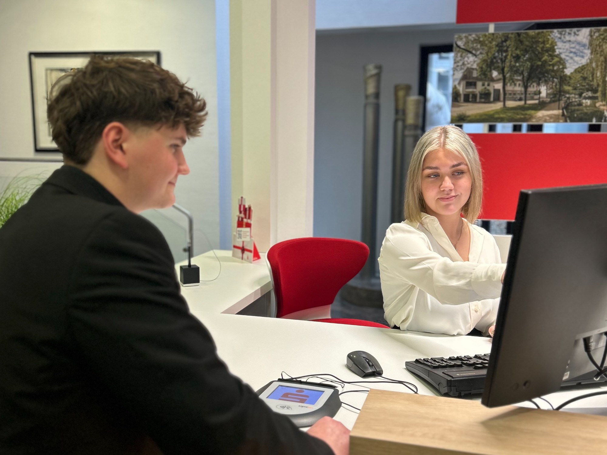 Drei junge Frauen sitzen gemeinsam an einem Schreibtisch mit Computerbildschirm in einem modernen Büro mit viel Grünpflanzen. Eine Frau tippt, die anderen hören interessiert zu.